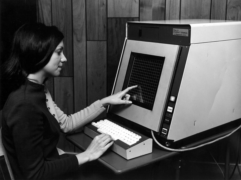 a woman touches what looks like an anchient computer screen with a graph paperlike surface. one of her hands is on the keyboard and the other is on the screen.